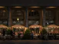 Terrasse de café animée à **heure de nuit**, éclairée par des lampes et des parasols rayés. Des clients discutent et boivent, profitant de l'ambiance parisienne.