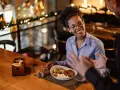 Une femme souriante discute avec un homme, son assiette de nourriture devant elle. Elle semble partager son avis sur la cuisine, une véritable critique culinaire.