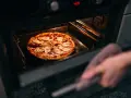 Une main avec une manique retire une pizza du four. La question est : quelle température pour réchauffer une pizza au four ?