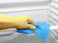 Main tenant, une main gantée de jaune nettoie les étagères d'un réfrigérateur avec un chiffon bleu. C'est le moment de nettoyer le condenseur du frigo pour qu'il fonctionne mieux.