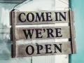 Texte annonce ouverture restaurant : panneau en bois "COME IN WE'RE OPEN" accroché à une porte vitrée.