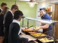 Des élèves font la queue pour le déjeuner, attendant leur plat. Le cuisinier, souriant, sert des légumes. Un contrat d'usage est peut-être en jeu pour ce repas scolaire.