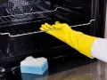 Main tenant porte-gants jaunes pour nettoyer le four. Une éponge bleue et blanche est posée sur le fond du four.