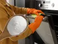 Une cuillère de bicarbonate de soude, le produit idéal avec quel produit nettoyer un four. Mains gantées nettoyant la porte du four.