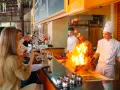 Cuisine ouverte d'un restaurant, chefs en action avec des flammes impressionnantes. Des clients prennent des photos, respectant la norme.