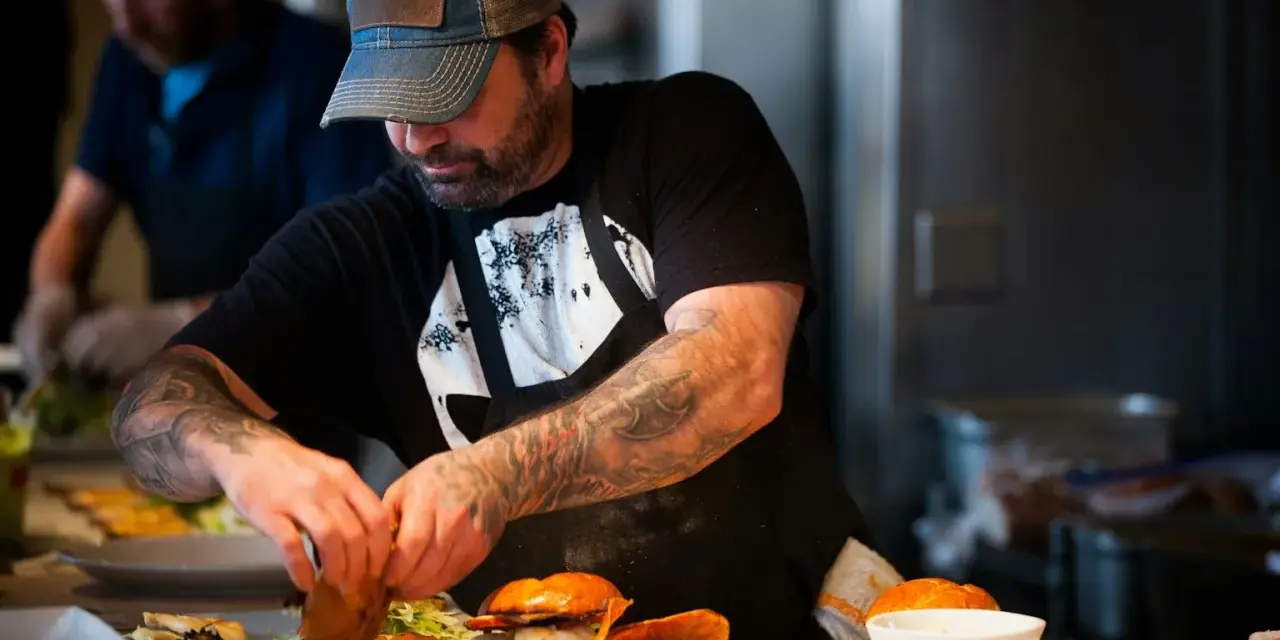 Un chef tatoué prépare des burgers et des frites, gage d'assurance restauration rapide.