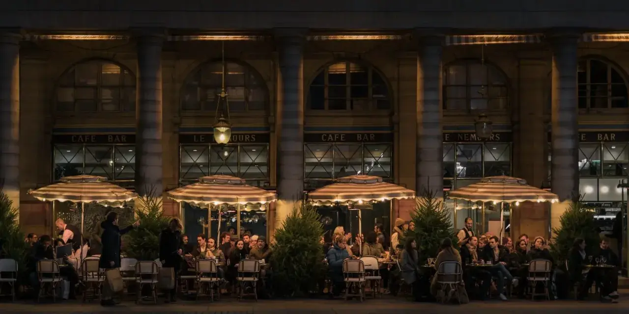 Terrasse de café animée à **heure de nuit**, éclairée par des lampes et des parasols rayés. Des clients discutent et boivent, profitant de l'ambiance parisienne.