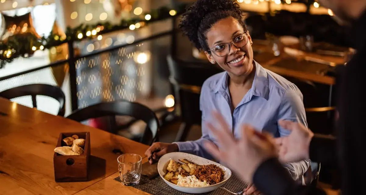 Une femme souriante discute avec un homme, son assiette de nourriture devant elle. Elle semble partager son avis sur la cuisine, une véritable critique culinaire.