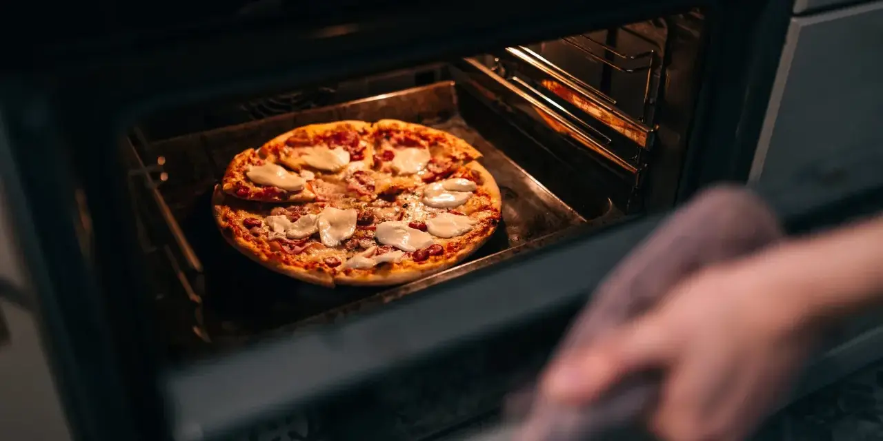 Une main avec une manique retire une pizza du four. La question est : quelle température pour réchauffer une pizza au four ?