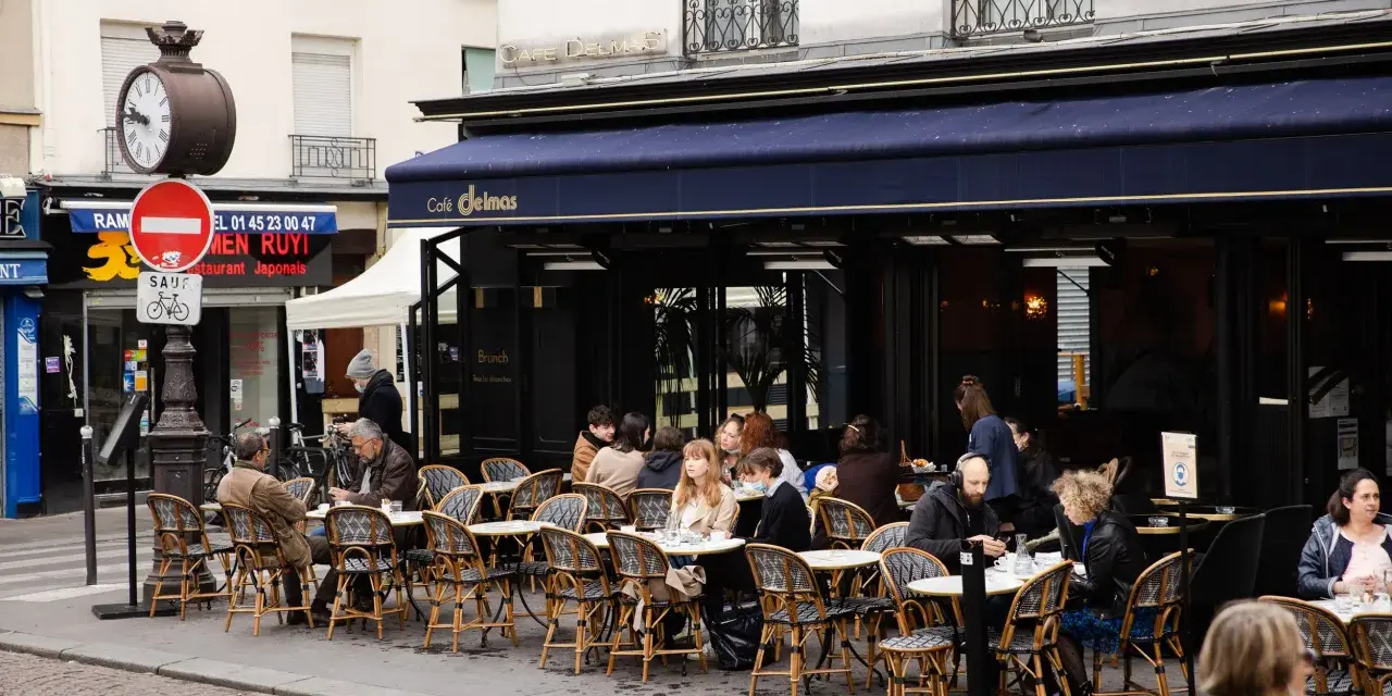 Terrasse animée d'un restaurant parisien, le Café Delmas, où des clients profitent de l'extérieur. Pas de tapage nocturne ici, juste une ambiance de bistrot paisible.