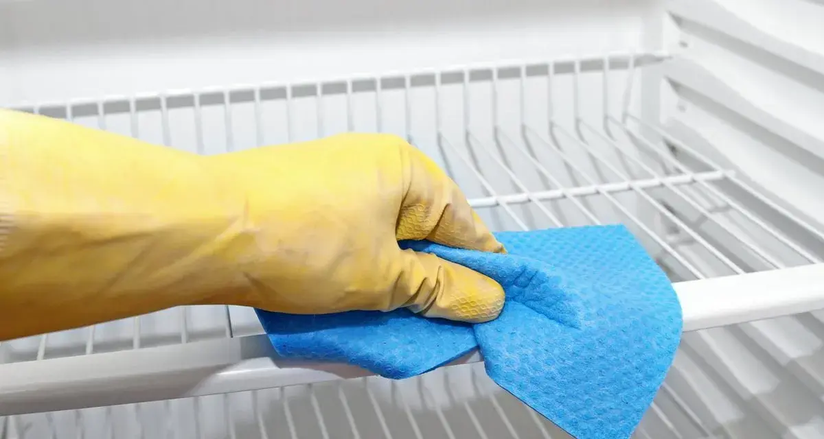 Main tenant, une main gantée de jaune nettoie les étagères d'un réfrigérateur avec un chiffon bleu. C'est le moment de nettoyer le condenseur du frigo pour qu'il fonctionne mieux.