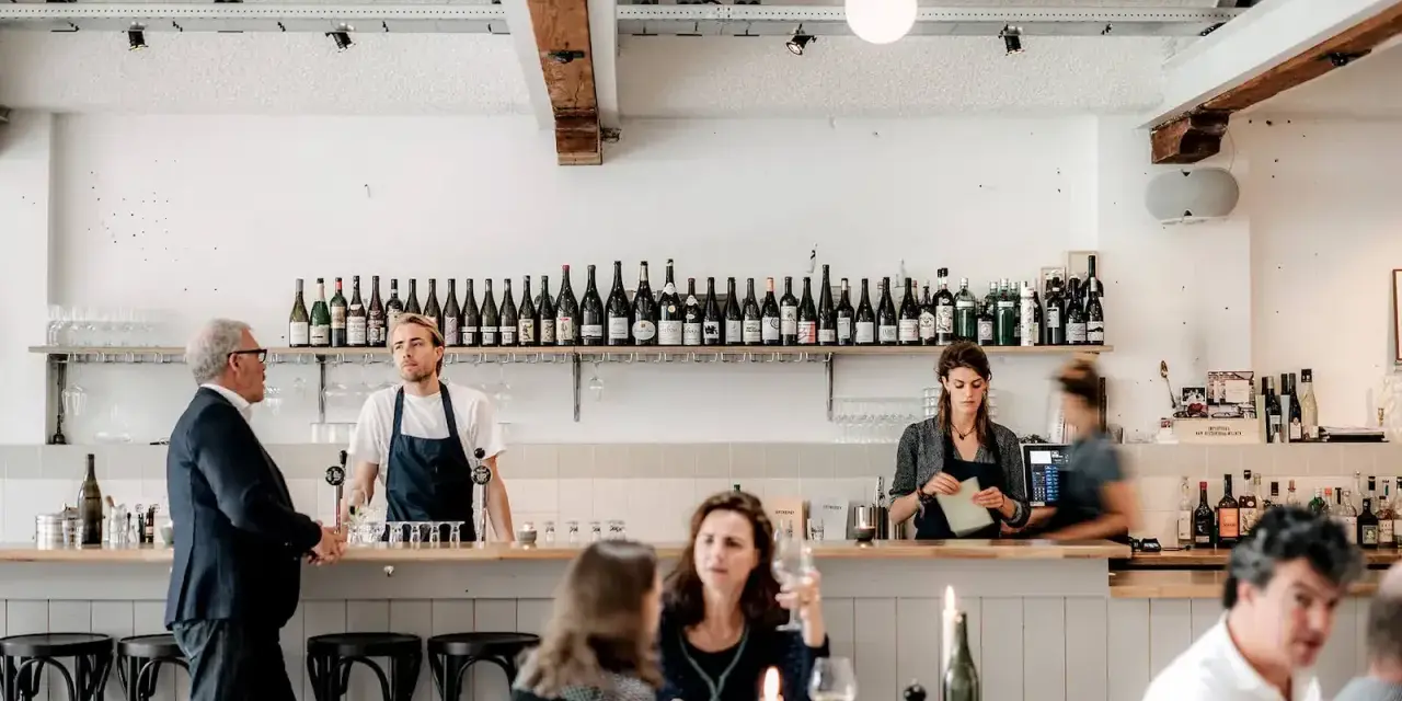 Un bar de restaurant animé avec un personnel attentif, des bouteilles de vin alignées et des clients profitant de l'ambiance. La gestion du restaurant assure un service fluide.