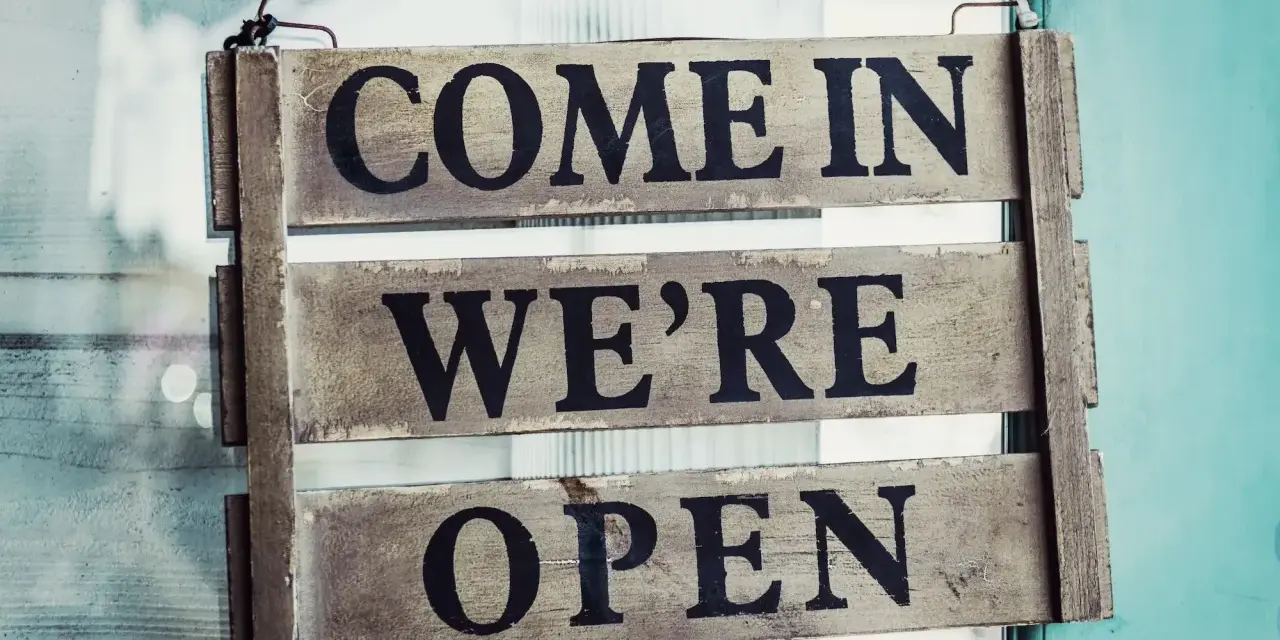 Texte annonce ouverture restaurant : panneau en bois "COME IN WE'RE OPEN" accroché à une porte vitrée.