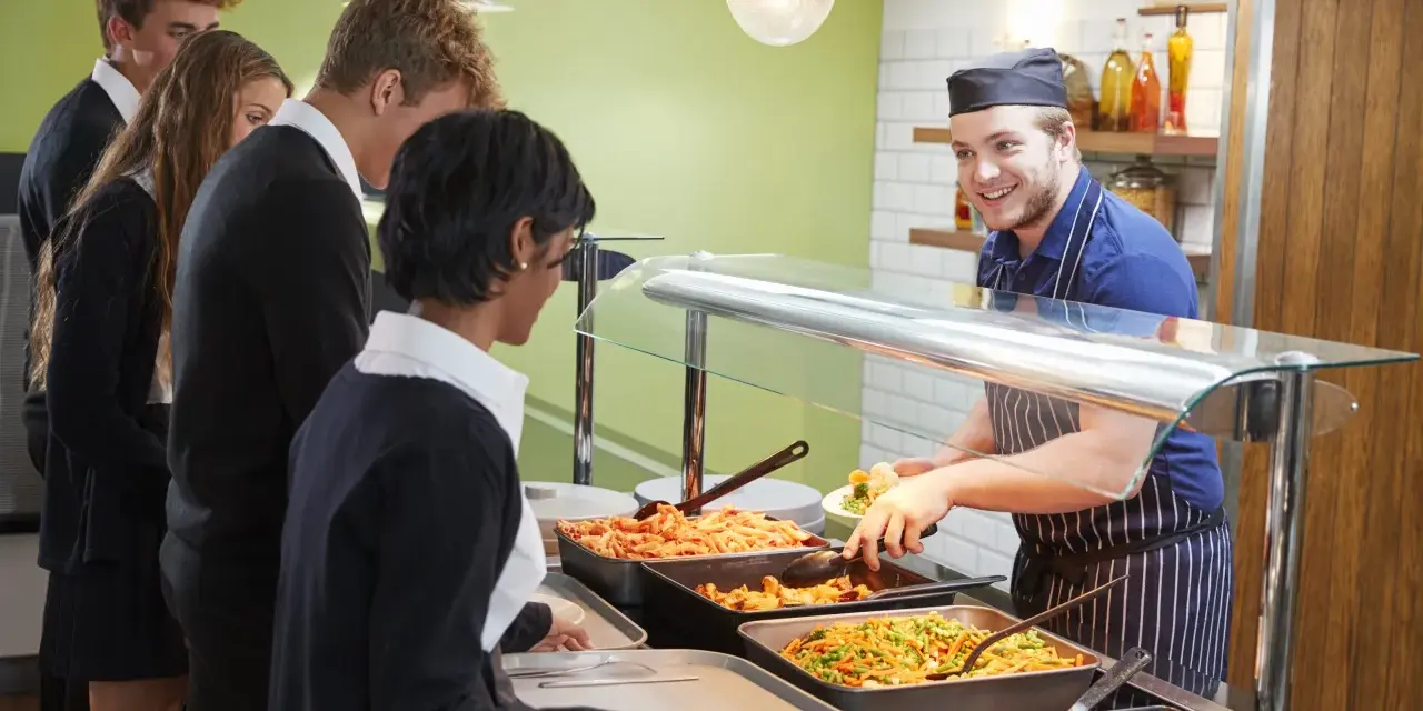 Des élèves font la queue pour le déjeuner, attendant leur plat. Le cuisinier, souriant, sert des légumes. Un contrat d'usage est peut-être en jeu pour ce repas scolaire.