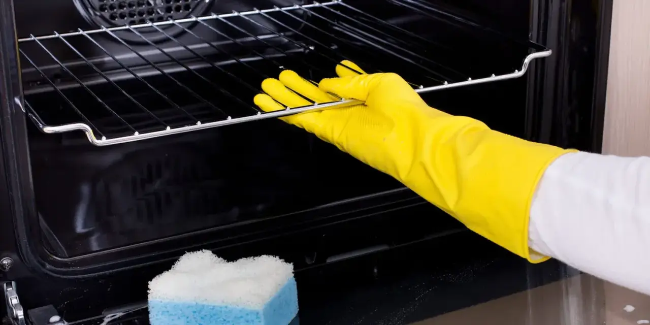 Main tenant porte-gants jaunes pour nettoyer le four. Une éponge bleue et blanche est posée sur le fond du four.