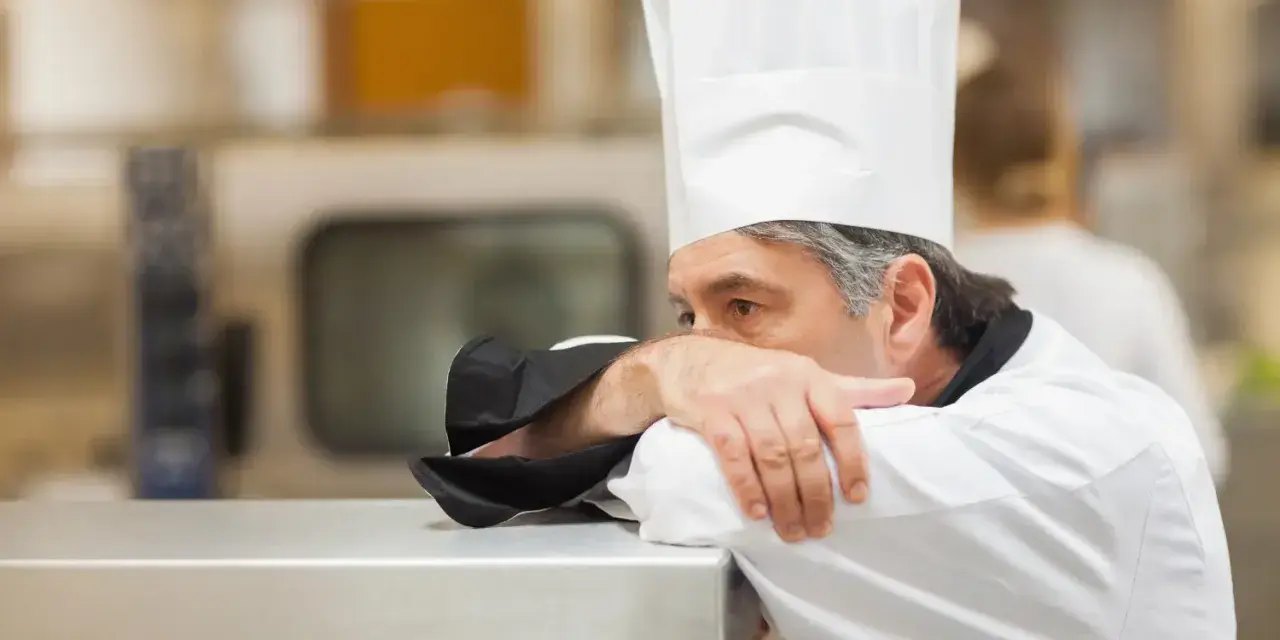 Un chef fatigué, le visage caché dans son bras, semble épuisé par un long jour de carence restauration rapide.