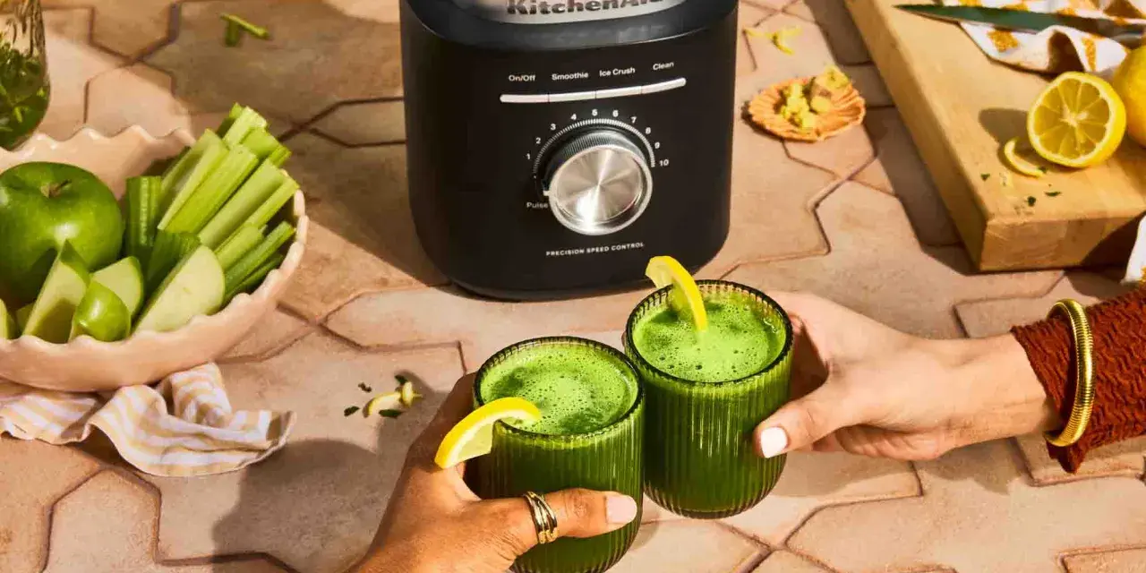 Deux mains portent des verres de jus vert, préparé avec un blender KitchenAid. Pommes, céleri et citrons sont prêts à être mixés.