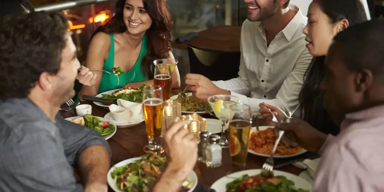 Un groupe d'amis partage un repas animé dans un restaurant, une excellente idée soirée à thème restaurant. Ils rient et discutent autour d'une table garnie de plats et de boissons.