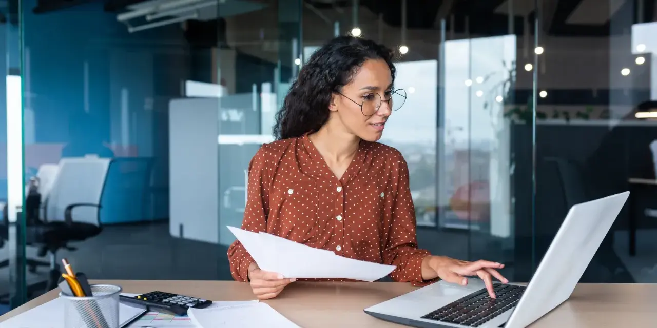 Femme travaillant sur un ordinateur portable, tenant des documents. Elle semble réfléchir à une décision unilatérale de l'employeur.