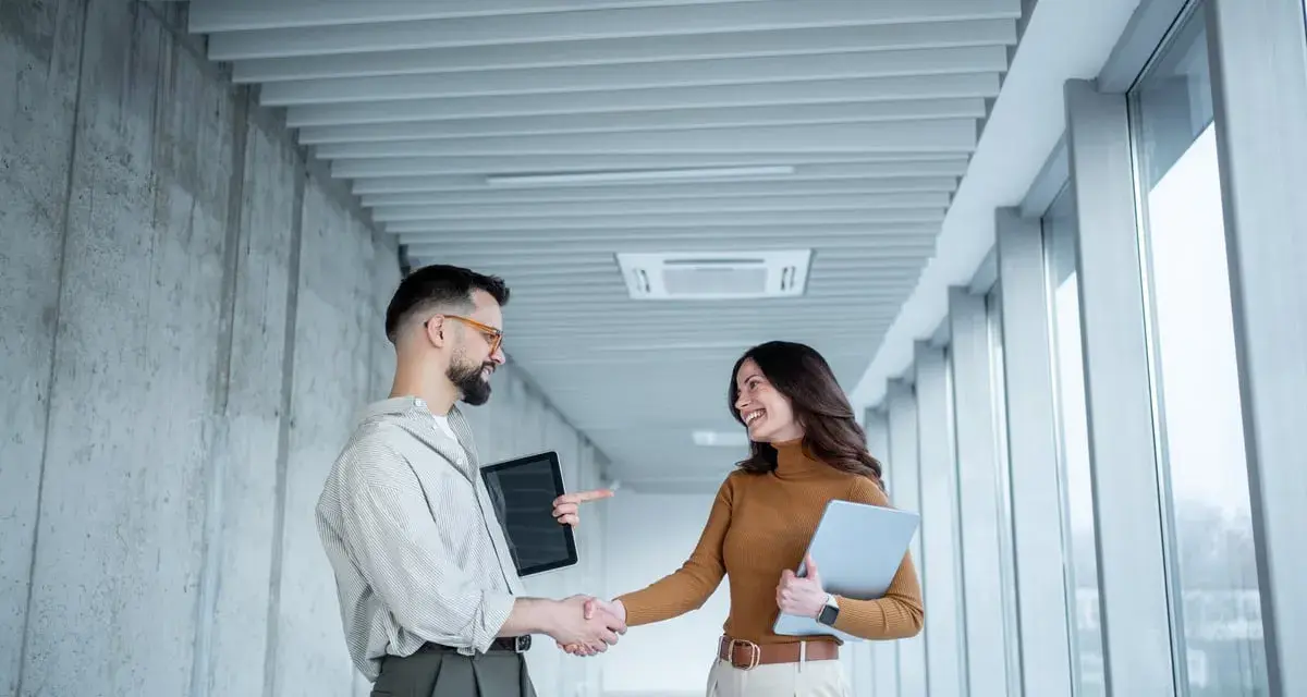 Un homme et une femme se serrent la main dans un couloir moderne, signe d'une prime cooptation réussie.