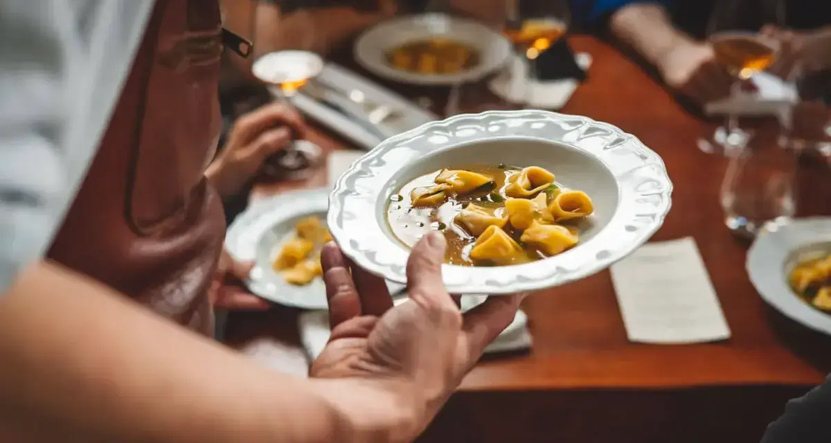 Un serveur présente une assiette de tortellinis dans un bouillon. Le service en restauration est à son apogée.