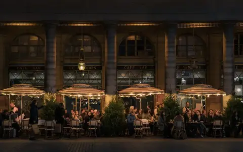 Terrasse de café animée à **heure de nuit**, éclairée par des lampes et des parasols rayés. Des clients discutent et boivent, profitant de l'ambiance parisienne.
