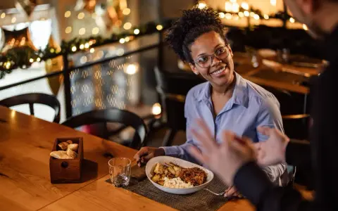 Une femme souriante discute avec un homme, son assiette de nourriture devant elle. Elle semble partager son avis sur la cuisine, une véritable critique culinaire.