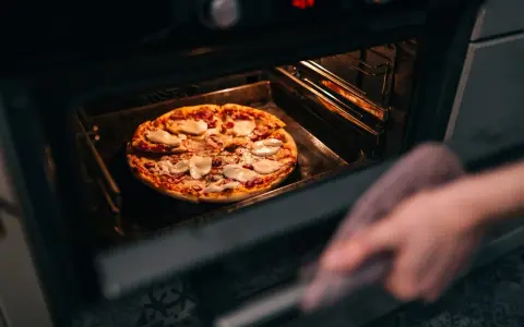 Une main avec une manique retire une pizza du four. La question est : quelle température pour réchauffer une pizza au four ?
