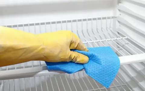 Main tenant, une main gantée de jaune nettoie les étagères d'un réfrigérateur avec un chiffon bleu. C'est le moment de nettoyer le condenseur du frigo pour qu'il fonctionne mieux.