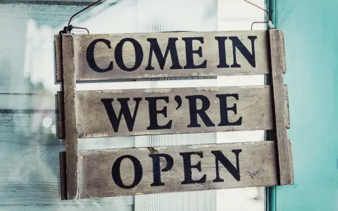 Texte annonce ouverture restaurant : panneau en bois "COME IN WE'RE OPEN" accroché à une porte vitrée.