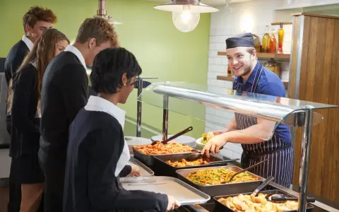 Des élèves font la queue pour le déjeuner, attendant leur plat. Le cuisinier, souriant, sert des légumes. Un contrat d'usage est peut-être en jeu pour ce repas scolaire.