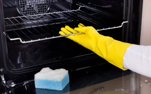 Main tenant porte-gants jaunes pour nettoyer le four. Une éponge bleue et blanche est posée sur le fond du four.