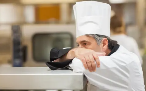 Un chef fatigué, le visage caché dans son bras, semble épuisé par un long jour de carence restauration rapide.