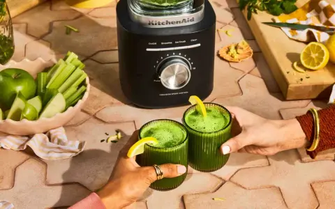Deux mains portent des verres de jus vert, préparé avec un blender KitchenAid. Pommes, céleri et citrons sont prêts à être mixés.