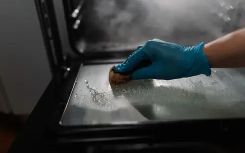 Main gantée bleue pour nettoyer le four avec du bicarbonate. La vapeur s'échappe, signe que le bicarbonate a chauffé.