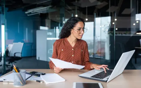 Femme travaillant sur un ordinateur portable, tenant des documents. Elle semble réfléchir à une décision unilatérale de l'employeur.