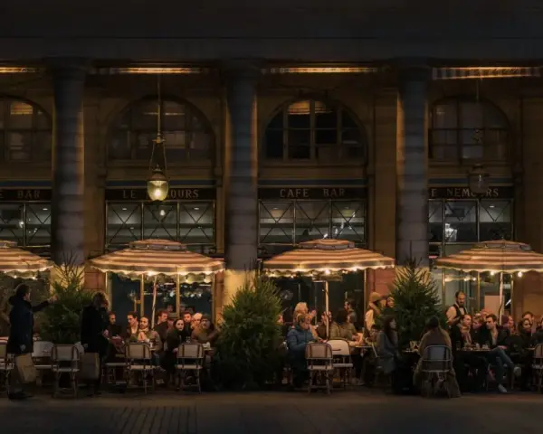 Terrasse de café animée à **heure de nuit**, éclairée par des lampes et des parasols rayés. Des clients discutent et boivent, profitant de l'ambiance parisienne.