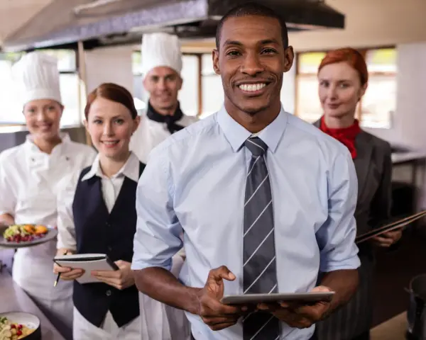 Une équipe souriante en cuisine, le responsable vérifie l'horaire collectif de travail sur sa tablette.