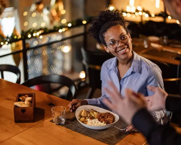 Une femme souriante discute avec un homme, son assiette de nourriture devant elle. Elle semble partager son avis sur la cuisine, une véritable critique culinaire.