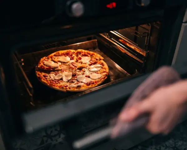 Une main avec une manique retire une pizza du four. La question est : quelle température pour réchauffer une pizza au four ?
