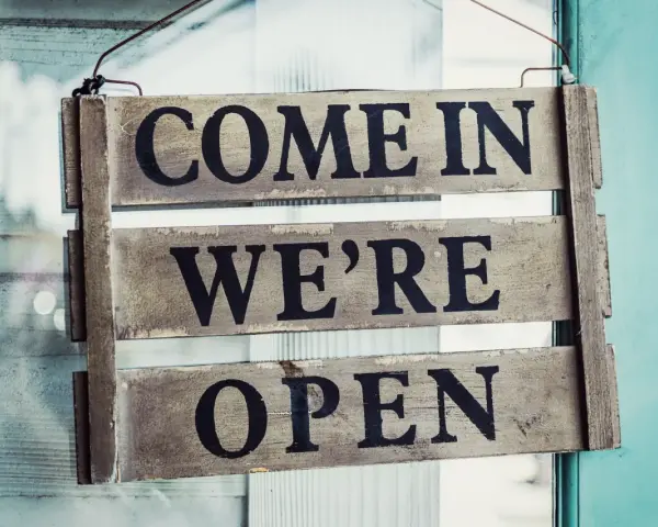 Texte annonce ouverture restaurant : panneau en bois "COME IN WE'RE OPEN" accroché à une porte vitrée.