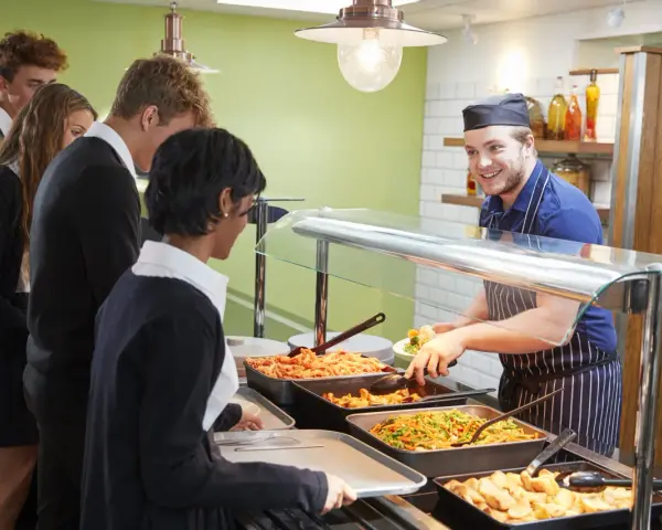 Des élèves font la queue pour le déjeuner, attendant leur plat. Le cuisinier, souriant, sert des légumes. Un contrat d'usage est peut-être en jeu pour ce repas scolaire.