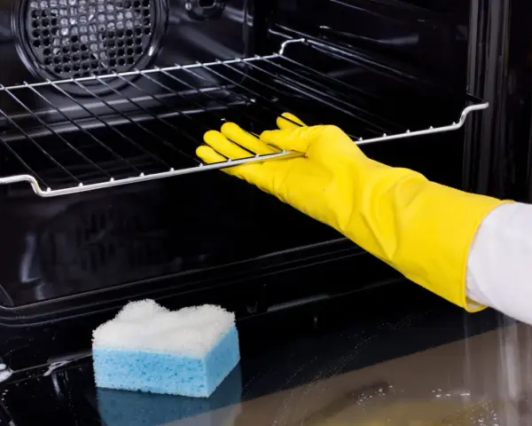 Main tenant porte-gants jaunes pour nettoyer le four. Une éponge bleue et blanche est posée sur le fond du four.