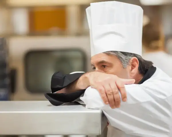Un chef fatigué, le visage caché dans son bras, semble épuisé par un long jour de carence restauration rapide.