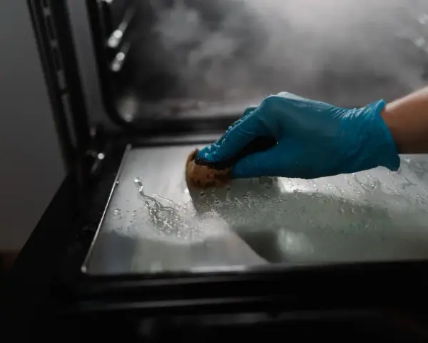 Main gantée bleue pour nettoyer le four avec du bicarbonate. La vapeur s'échappe, signe que le bicarbonate a chauffé.