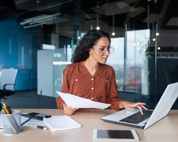 Femme travaillant sur un ordinateur portable, tenant des documents. Elle semble réfléchir à une décision unilatérale de l'employeur.