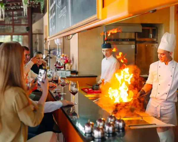 Cuisine ouverte d'un restaurant, chefs en action avec des flammes impressionnantes. Des clients prennent des photos, respectant la norme.