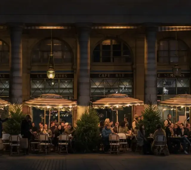Terrasse de café animée à **heure de nuit**, éclairée par des lampes et des parasols rayés. Des clients discutent et boivent, profitant de l'ambiance parisienne.