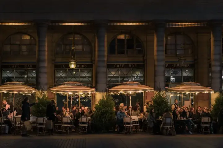 Terrasse de café animée à **heure de nuit**, éclairée par des lampes et des parasols rayés. Des clients discutent et boivent, profitant de l'ambiance parisienne.