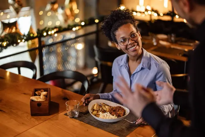 Une femme souriante discute avec un homme, son assiette de nourriture devant elle. Elle semble partager son avis sur la cuisine, une véritable critique culinaire.